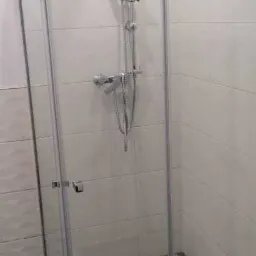 Kabina prysznicowa z chromowaną armaturą, wykończona białą płytką i mozaikową podłogą z odpływem liniowym. W górnej części ściany dekoracyjny pas z mozaiki.