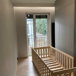 good - Jasne wnętrze pokoju z drewnianą podłogą w jodełkę, łóżeczkiem dziecięcym z surowego drewna ustawionym przy oknie balkonowym z widokiem na zieleń, minimalistyczna lampa sufitowa i szara szafa w tle...