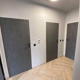 good - Nowoczesne, szare drzwi wewnętrzne z czarnymi klamkami, zamontowane w jasnym korytarzu z parkietem ułożonym w jodełkę.