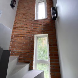 good - Klatka schodowa z szarymi schodami i ścianą z czerwonej cegły z dwoma oknami, oświetlona czarnymi kinkietami.