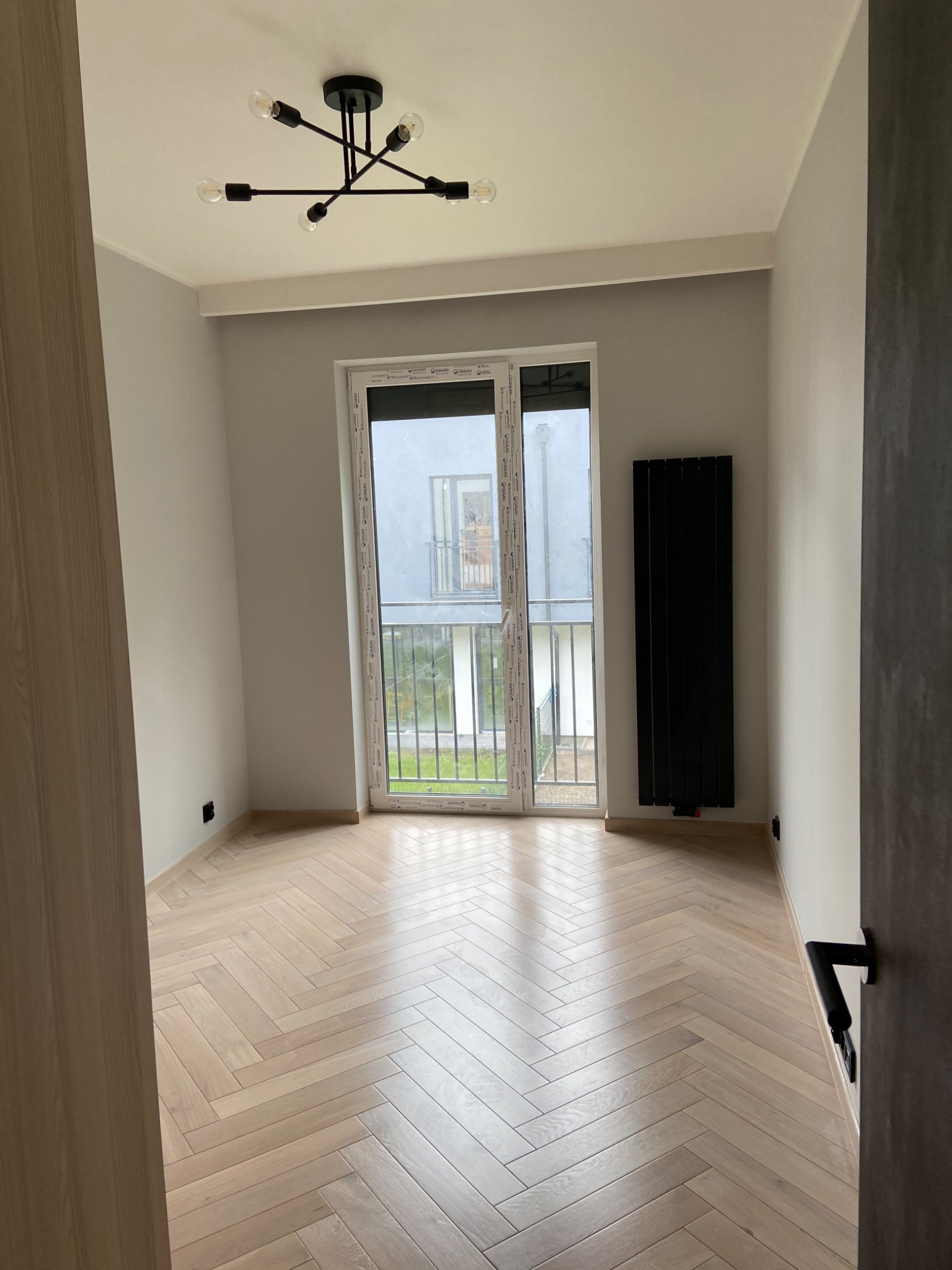 Jasny parkiet ułożony we wzór jodełki w pustym pokoju z szarymi ścianami, czarnym grzejnikiem i nowoczesną lampą sufitową.