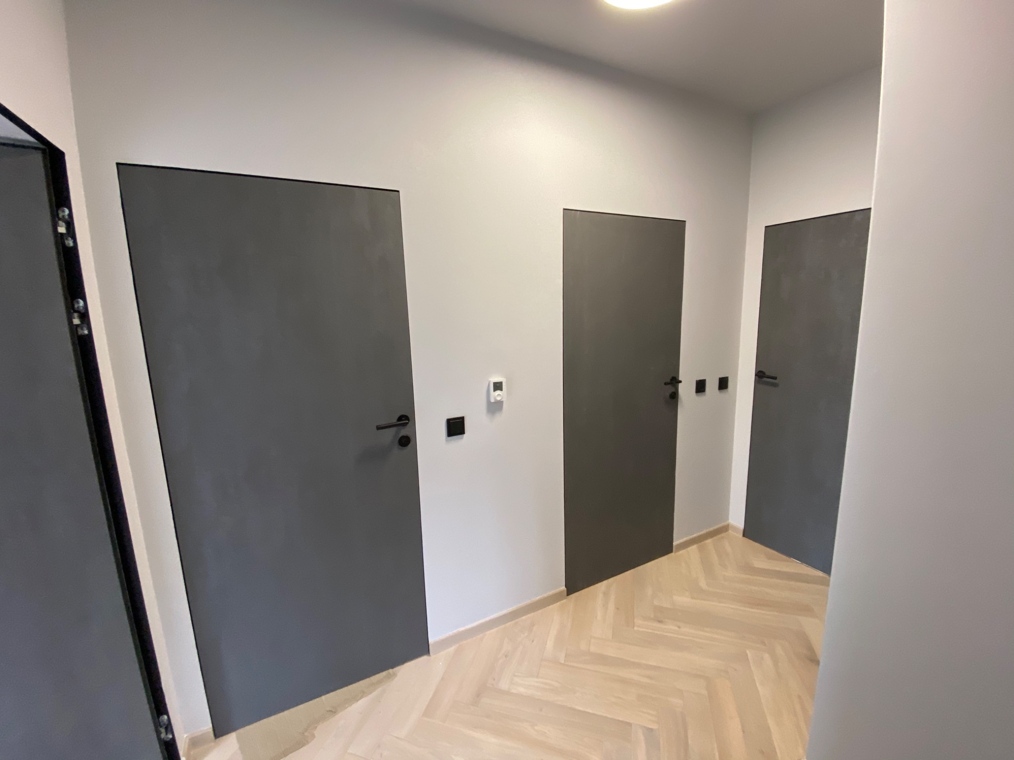 Nowoczesne, szare drzwi wewnętrzne z czarnymi klamkami, zamontowane w jasnym korytarzu z parkietem ułożonym w jodełkę.