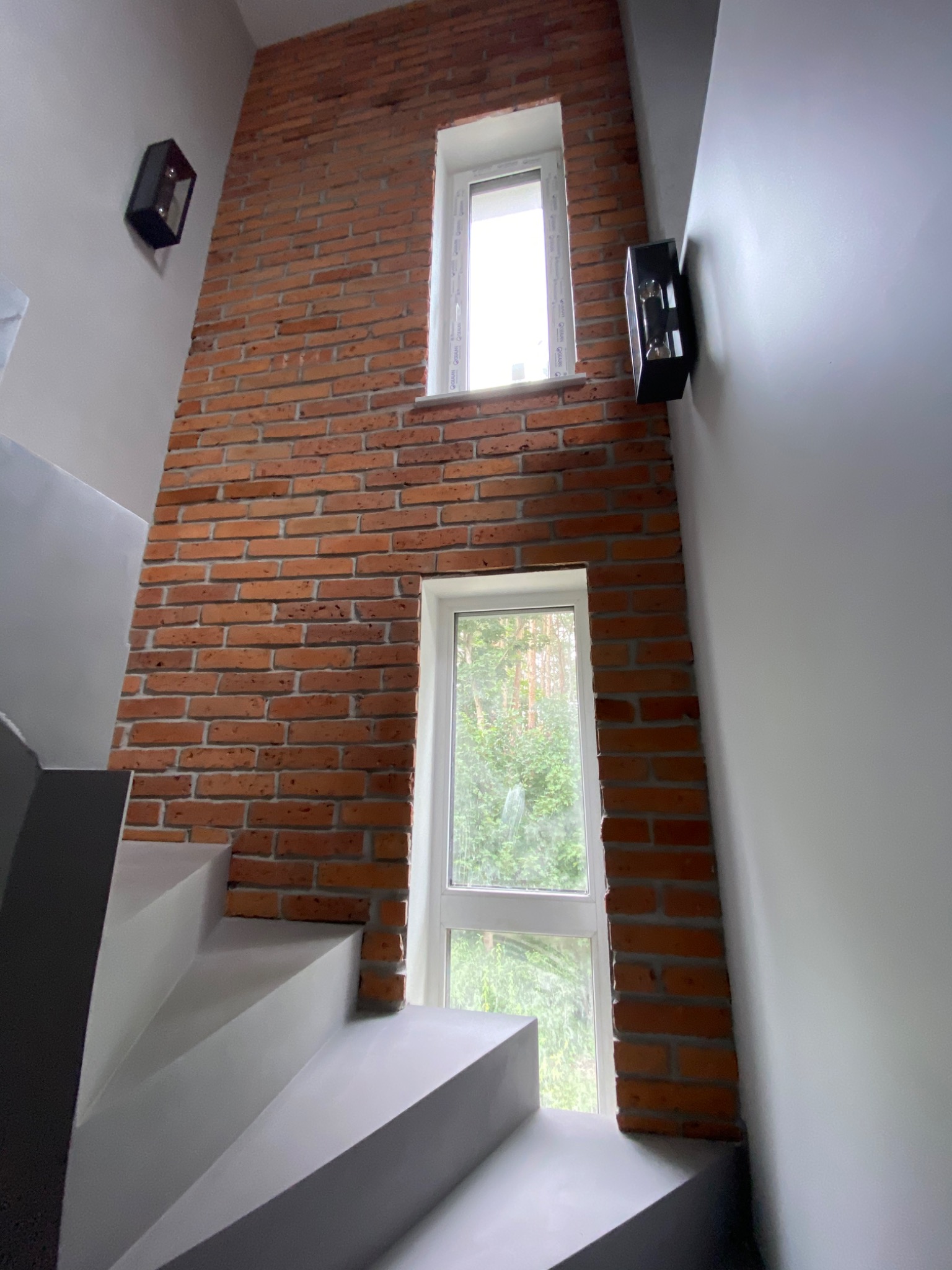 Klatka schodowa z szarymi schodami i ścianą z czerwonej cegły z dwoma oknami, oświetlona czarnymi kinkietami.