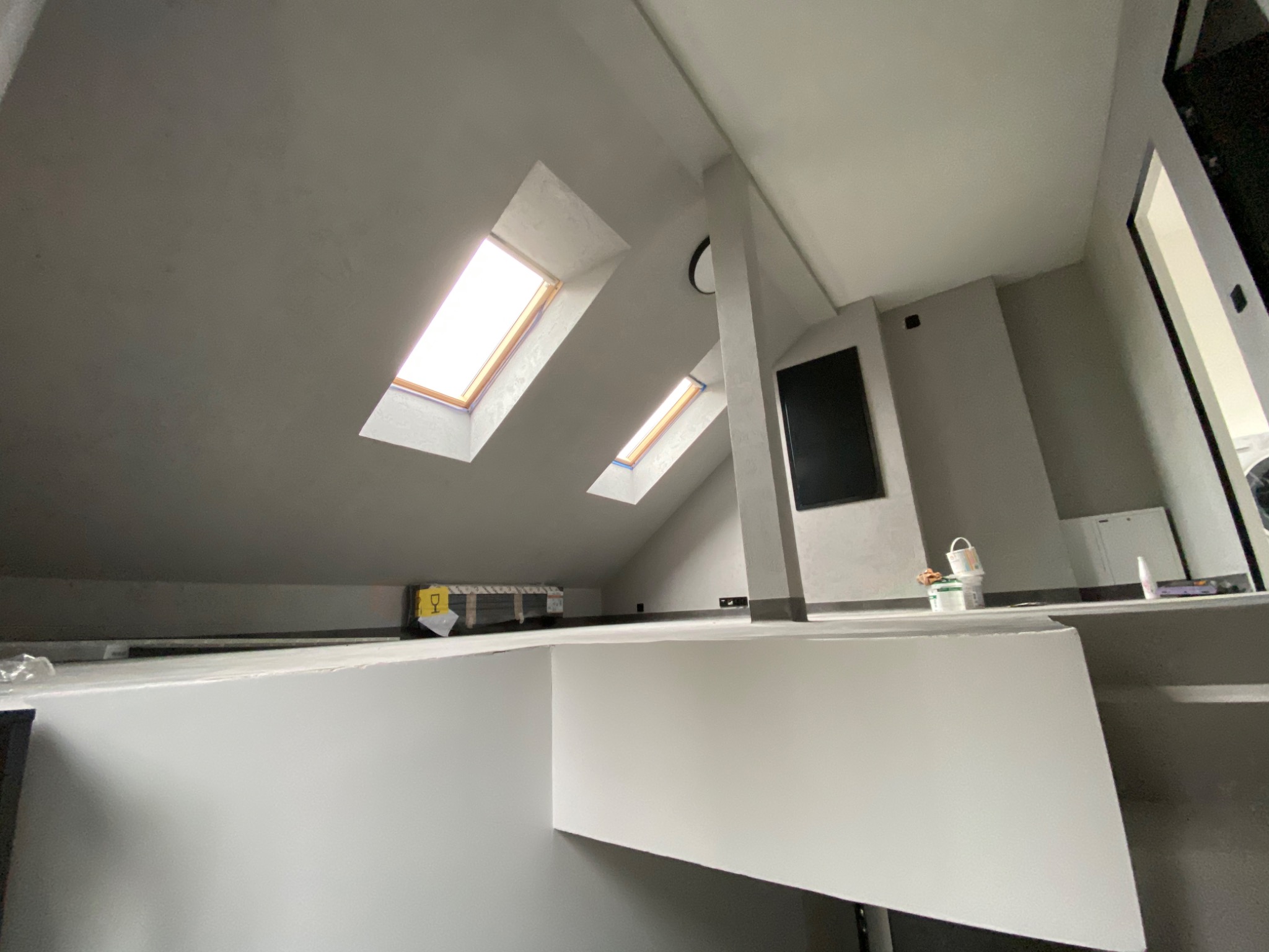 Poddasze po szpachlowaniu i malowaniu na szaro, widoczne dwa okna dachowe, kolumna konstrukcyjna, oraz przygotowane materiały malarskie.