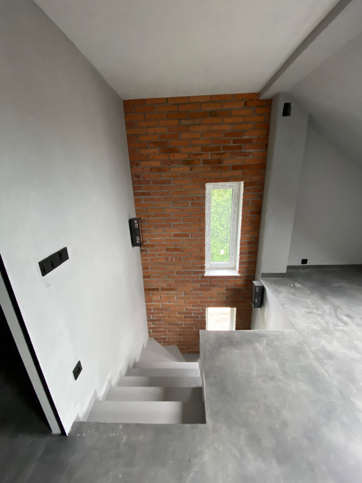 Wnętrze domu w trakcie wykańczania: schody pomalowane na szaro, ściana z czerwonej cegły z oknem, fragment podłogi z wylewki samopoziomującej, widoczne puszki elektryczne.