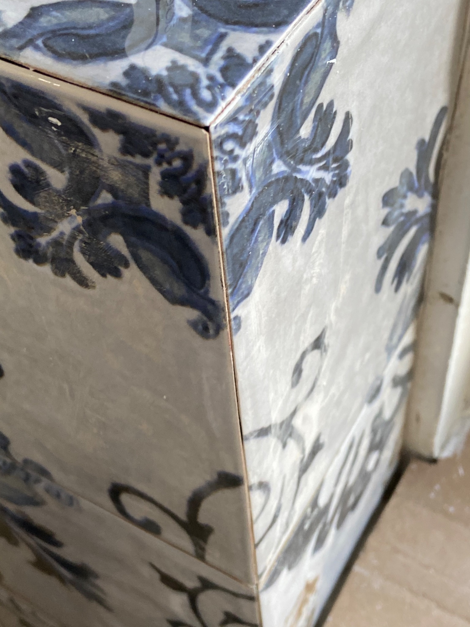 Fragment narożnika obłożonego białymi płytkami z niebieskim, kwiatowym wzorem, widoczne delikatne fugi i niedoskonałości wykończenia.