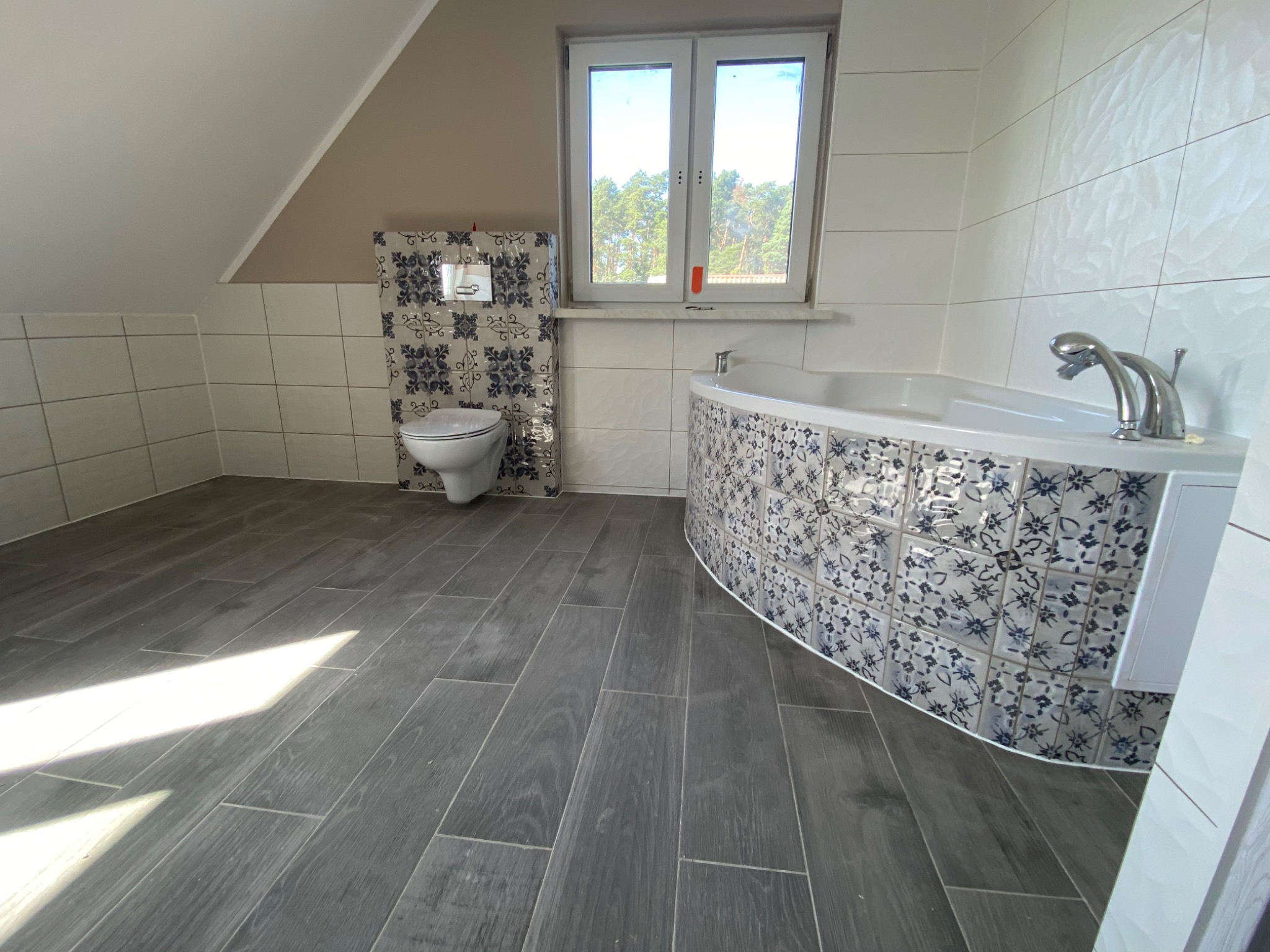 Nowoczesna łazienka na poddaszu z szarą podłogą imitującą drewno, białe płytki ścienne z delikatną fakturą, toaleta i wanna obudowane płytkami w niebiesko-białe wzory, okno z widokiem na drzewa.