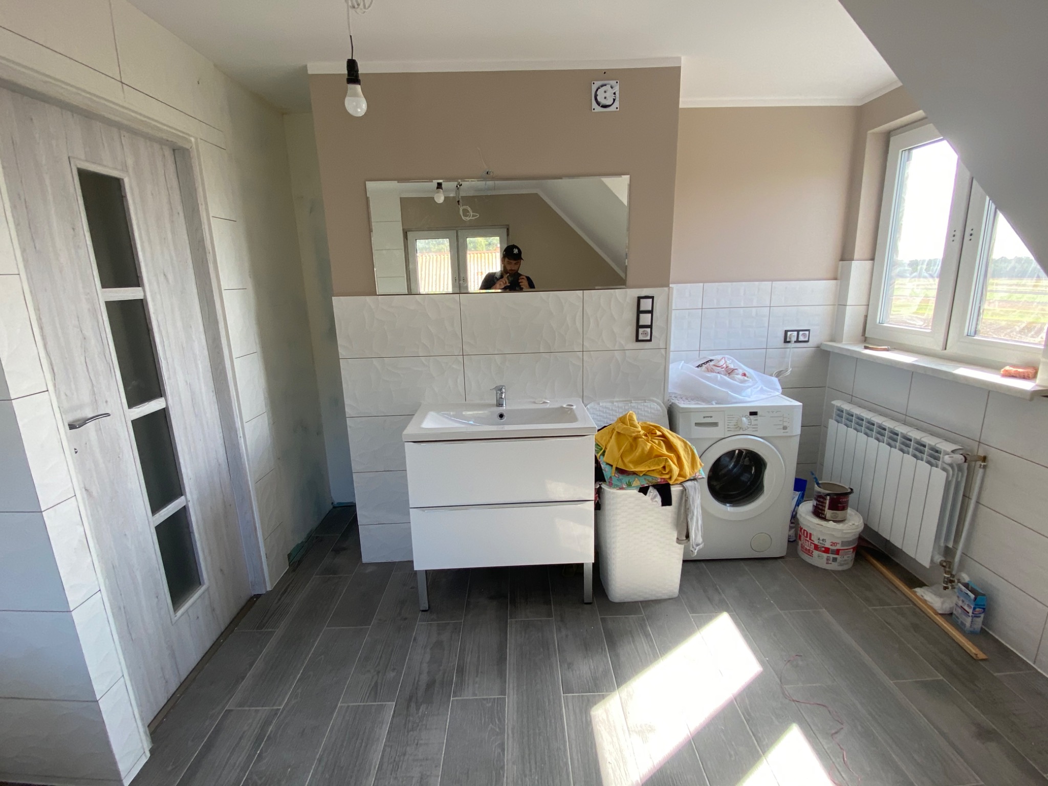 Wykończona łazienka na poddaszu z szarymi płytkami podłogowymi imitującymi drewno, białą umywalką z szafką, pralką, grzejnikiem i lustrem, w którym odbija się osoba robiąca zdjęcie.