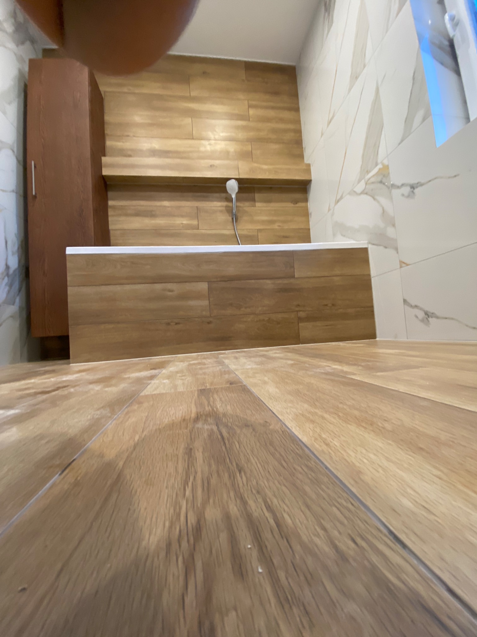 Nowoczesna łazienka z wykończeniem w odcieniach drewna i bieli, widok z niskiego kąta na podłogę w kierunku wanny obudowanej panelami imitującymi drewno, z widoczną kabiną prysznicową i szafką.