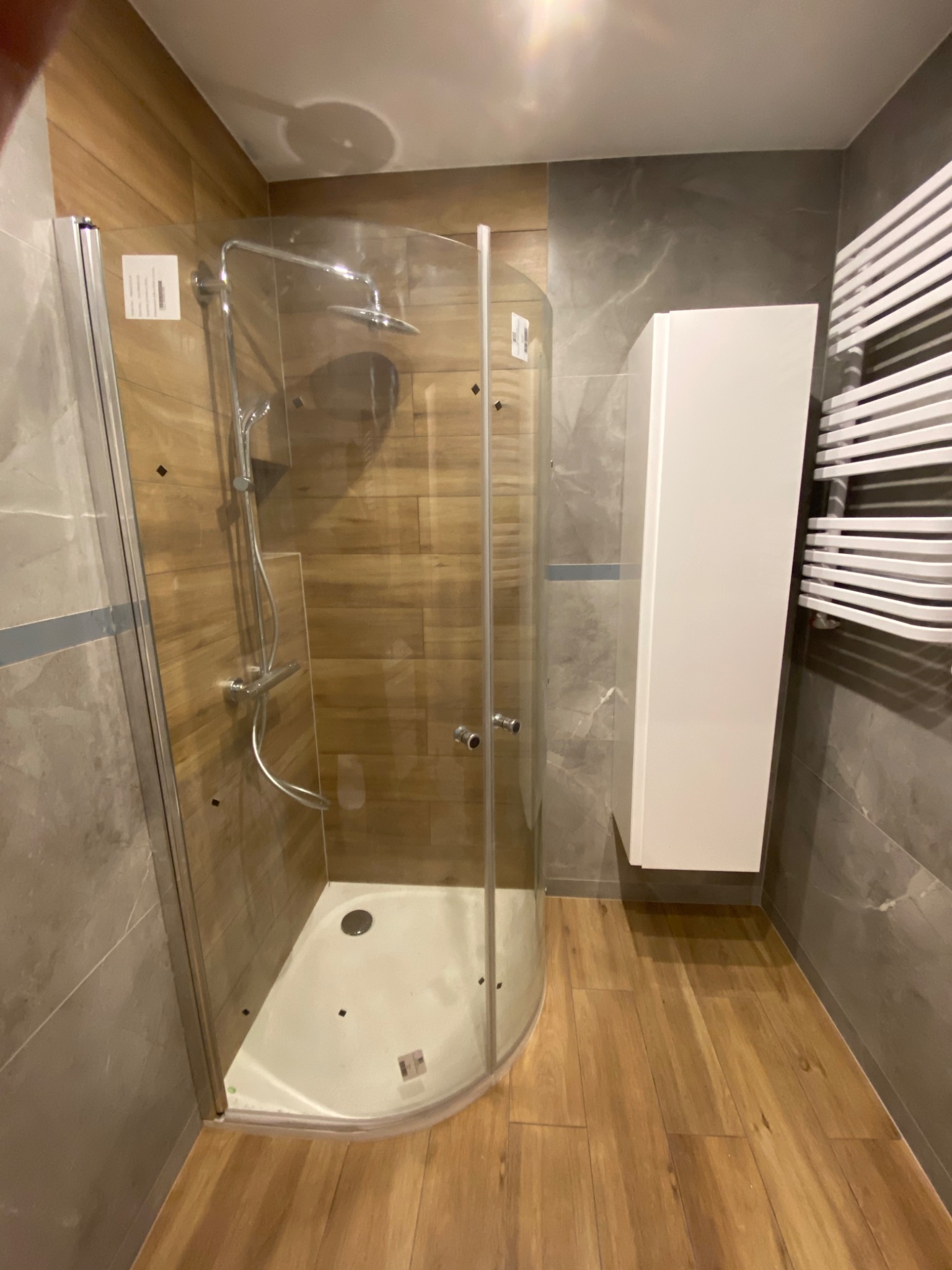 Kabina prysznicowa z zaokrąglonym szkłem, drewnopodobne płytki na ścianie wewnątrz kabiny, szare płytki imitujące marmur na pozostałych ścianach, biała szafka wisząca i grzejnik drabinkowy w łazience.