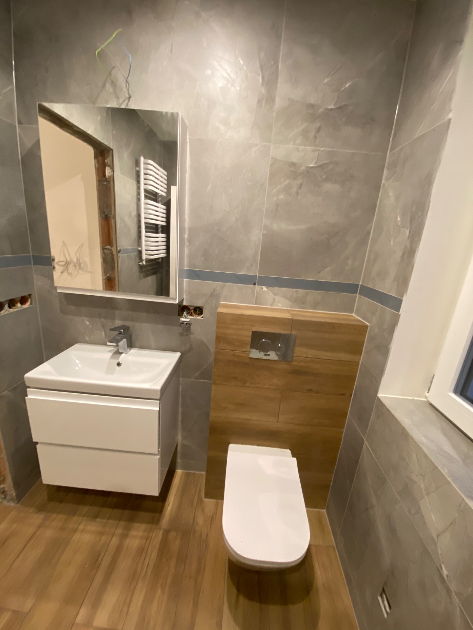 Nowoczesna łazienka w szarościach z drewnianymi akcentami: umywalka z szafką, toaleta podwieszana z drewnianą obudową, lustro z widocznym grzejnikiem w odbiciu, płytki imitujące marmur na ścianach,...