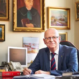 Portret mężczyzny w garniturze, z piórem w dłoni, piszącego w otwartym notatniku na biurku, w tle obrazy w złotych ramach i kalendarz.