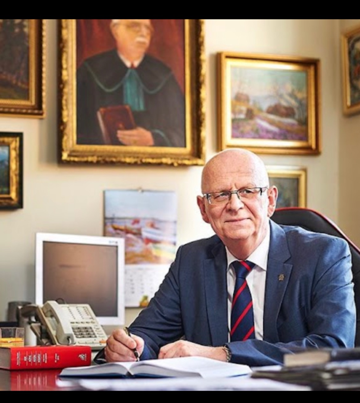 Portret mężczyzny w garniturze, z piórem w dłoni, piszącego w otwartym notatniku na biurku, w tle obrazy w złotych ramach i kalendarz.