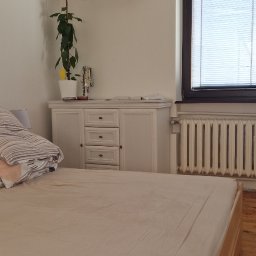 COOL PROJECT ŁUKASZ BIELAWSKI - Biały klimatyzator zamontowany nad komodą w sypialni, oświetlony dekoracyjnymi lampkami w kształcie kul, z widocznym fragmentem łóżka i grzejnika.