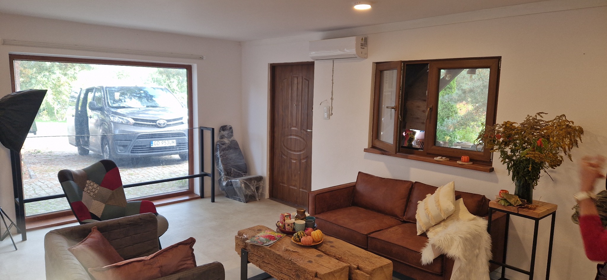 Wnętrze pokoju z klimatyzatorem ściennym nad drzwiami, brązowa sofa i drewniany stolik kawowy, widok na samochód przez okno.