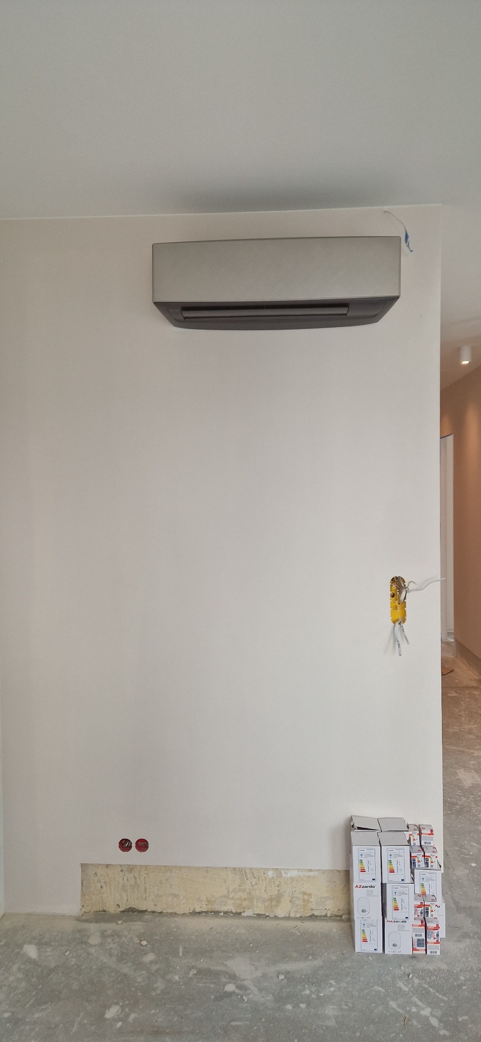 Zamontowana, srebrna jednostka klimatyzacji na białej ścianie w trakcie remontu, widoczne wystające kable elektryczne i stos kartonów na podłodze.