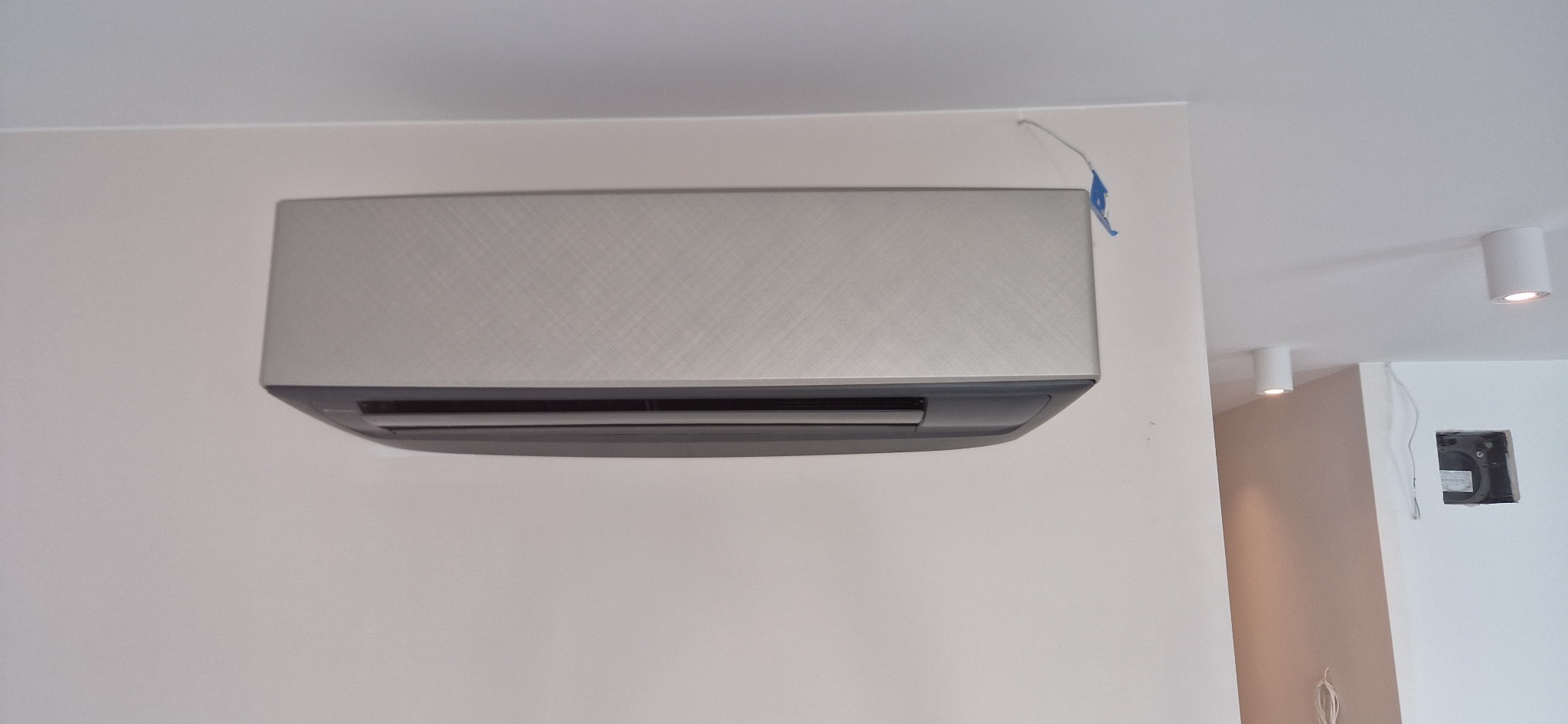Szary klimatyzator ścienny zamontowany w jasnym pomieszczeniu z widocznymi przewodami i punktowymi lampami sufitowymi.