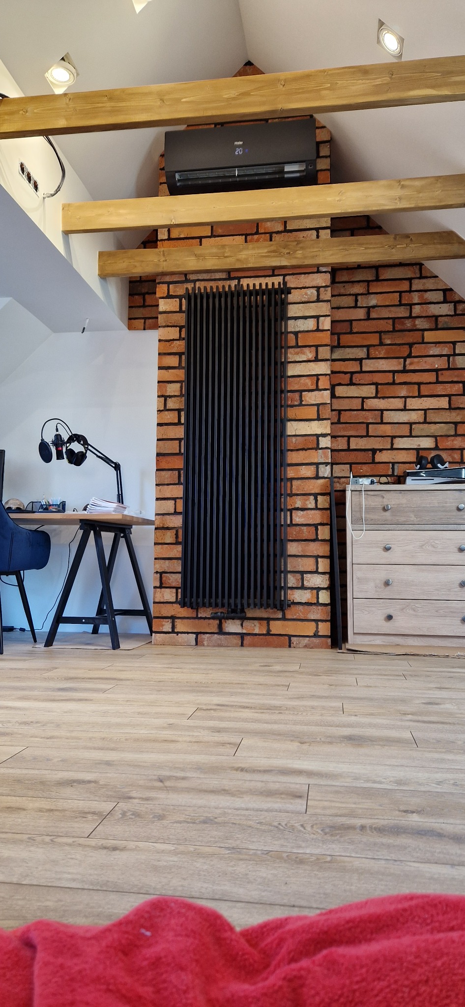 Czarny klimatyzator Haier zamontowany na ceglanej ścianie w domu, widoczny również czarny grzejnik panelowy, biurko z mikrofonem i drewniana komoda w pokoju z poddaszem.