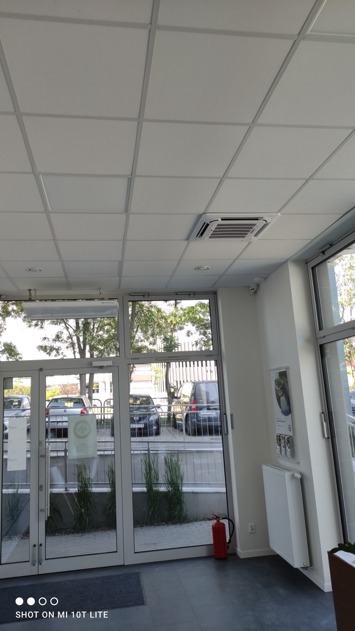 Kasetonowa jednostka klimatyzacyjna zamontowana w suficie podwieszanym w biurze, widoczna z perspektywy podłogi, z widokiem na szklane drzwi wejściowe i parking.