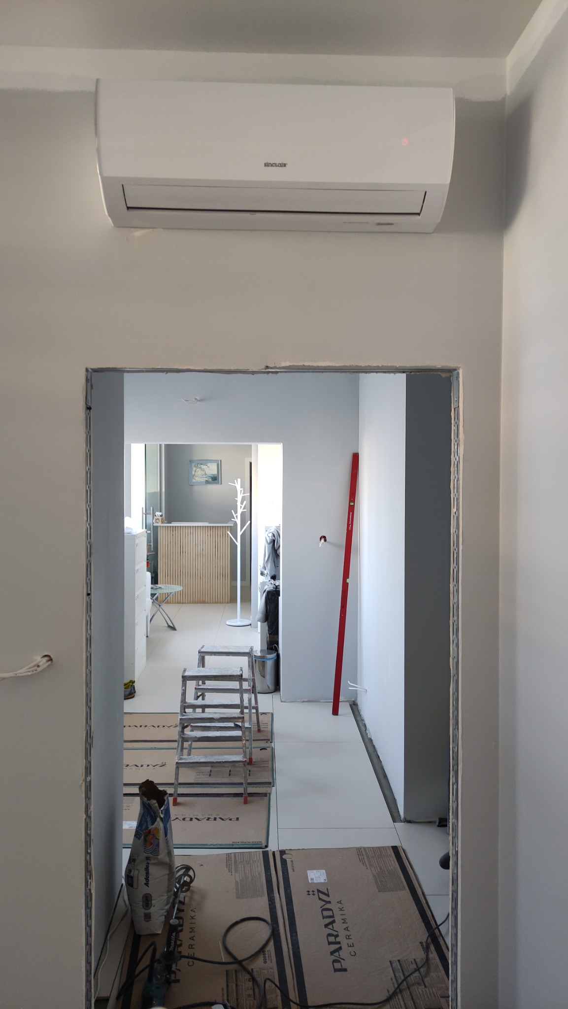Biała jednostka klimatyzacji Sinclair zamontowana na ścianie w jasnym pomieszczeniu, widoczna przez framugę drzwi w trakcie remontu, ze schodkami i workami z zaprawą na podłodze.