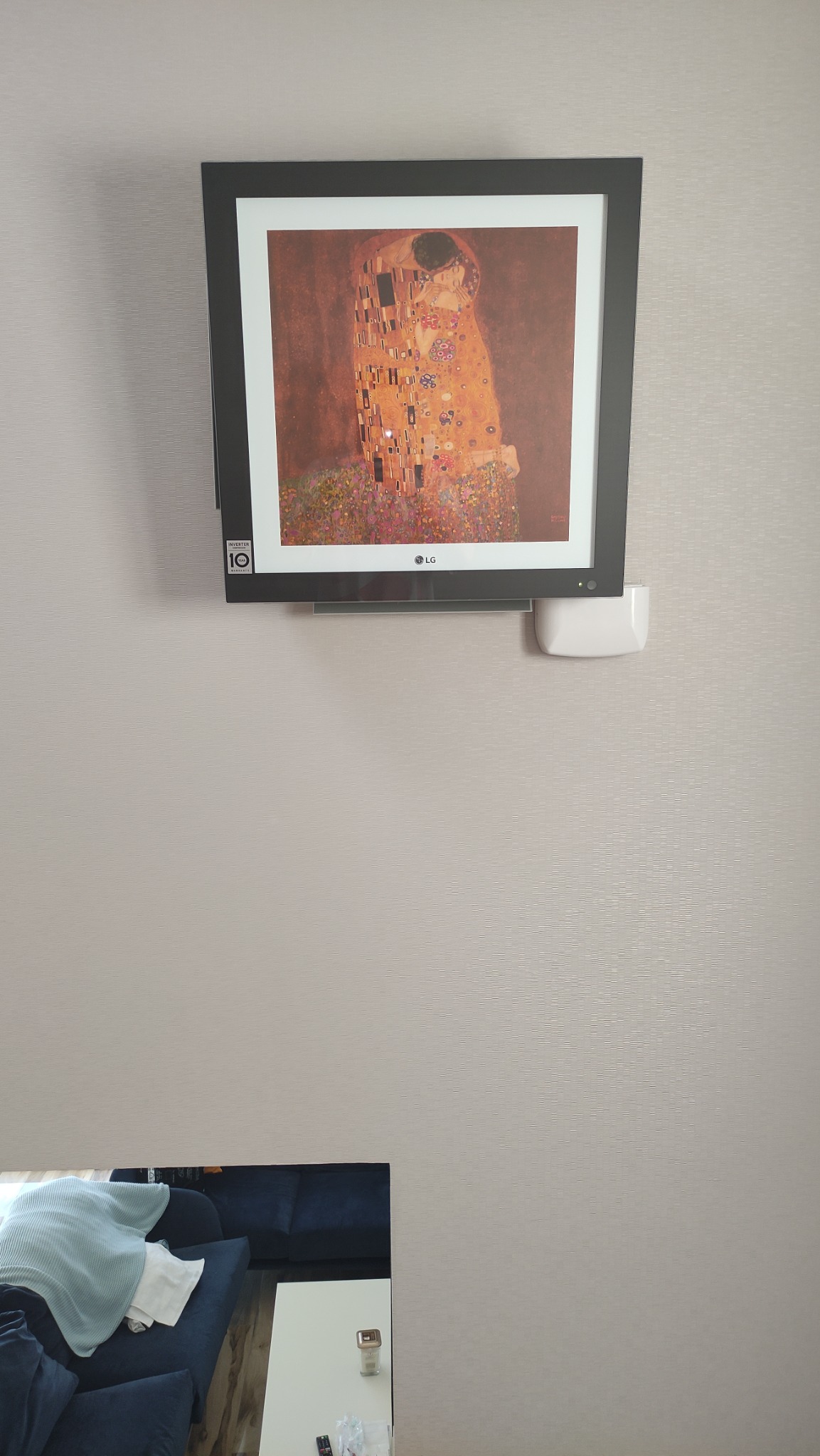 Obraz 'Pocałunek' Gustava Klimta w czarnej ramie, zamontowany na ścianie nad niebieską sofą i białym stolikiem kawowym. Widoczny biały element instalacji pod ramą obrazu.