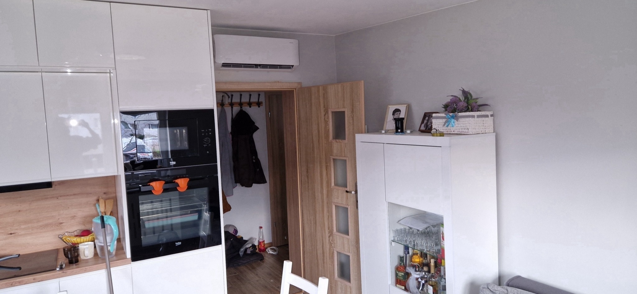 Biały klimatyzator ścienny zamontowany w nowoczesnym mieszkaniu nad drzwiami do garderoby w pobliżu zabudowy kuchennej.