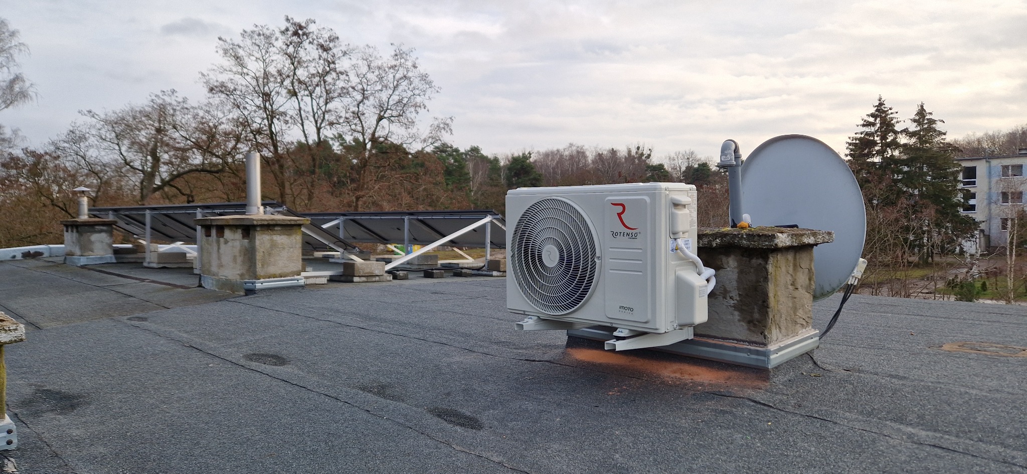 Biała jednostka zewnętrzna klimatyzacji marki Rotenso zamontowana na płaskim dachu budynku, obok widoczna antena satelitarna i panele słoneczne.