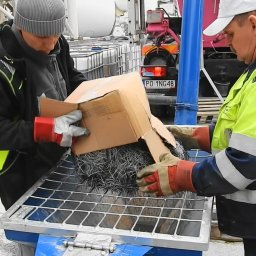 Dekor PUHB - Dwóch pracowników w odzieży roboczej wsypuje włókna stalowe z kartonowego pudełka na metalową kratę, w tle betoniarka i samochód z widoczną tablicą rejestracyjną.