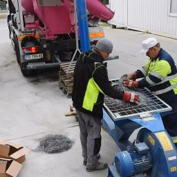 Dekor PUHB - Dwóch pracowników w roboczych ubraniach przygotowuje włókna stalowe do betonu na tle betonomieszarki i pojemników IBC