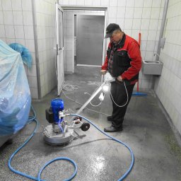 Dekor PUHB - Mężczyzna w czerwonej kurtce obsługuje maszynę do czyszczenia posadzki w pomieszczeniu wyłożonym białymi płytkami, widoczny niebieski wąż i kabel zasilający, podłoga mokra i częściowo zabrudzona.
