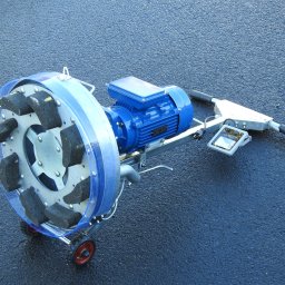 Dekor PUHB - Szlifierka do betonu i lastriko z niebieskim silnikiem elektrycznym, tarczą z segmentami ściernymi i przezroczystą osłoną, stojąca na mokrej, asfaltowej nawierzchni.