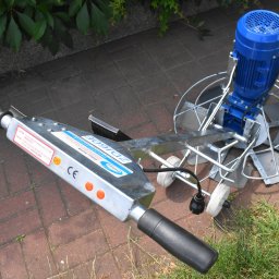 Dekor PUHB - Zacieraczka do betonu z niebieskim silnikiem elektrycznym, widoczna na kostce brukowej, z fragmentami trawy i liści w tle.