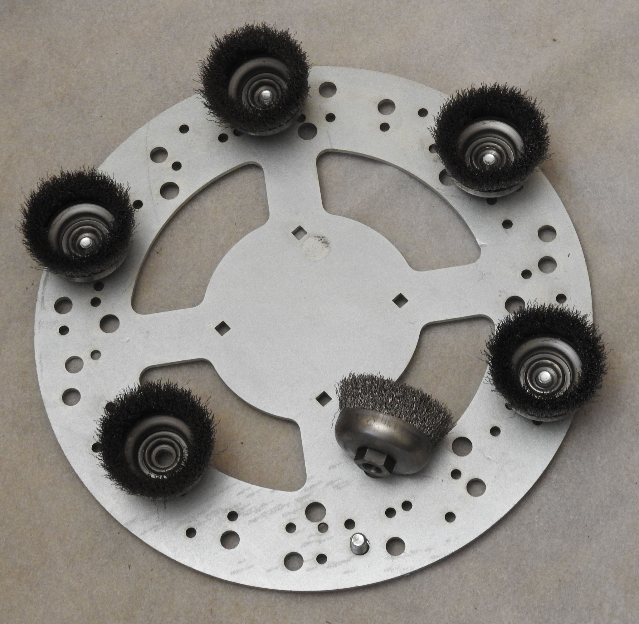 Tarcza szlifierska z sześcioma zamocowanymi, okrągłymi szczotkami drucianymi do czyszczenia powierzchni, jedna szczotka leży obok tarczy.