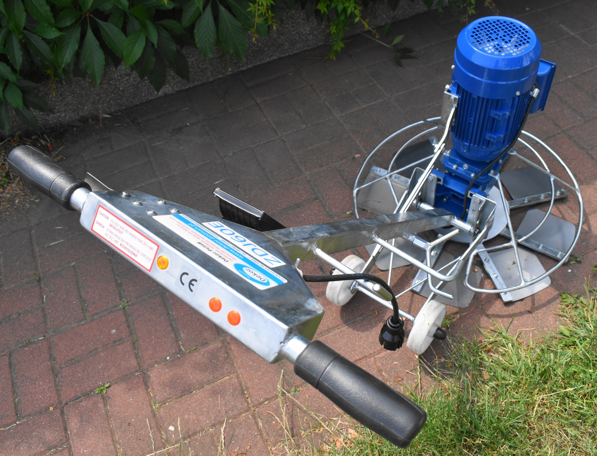 Zacieraczka do betonu z niebieskim silnikiem elektrycznym, widoczna na kostce brukowej, z fragmentami trawy i liści w tle.