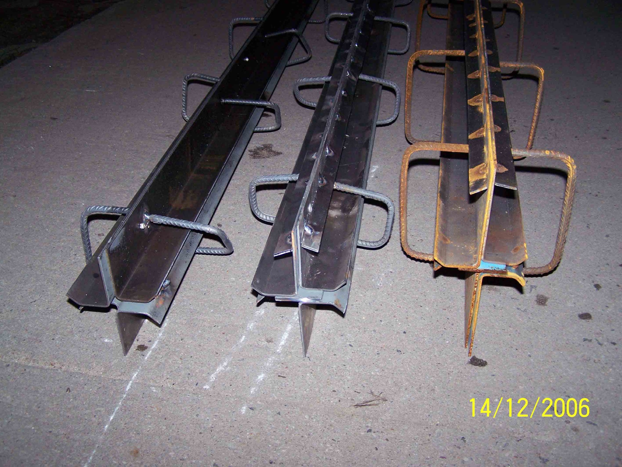 Trzy metalowe profile z ostrymi zakończeniami i uchwytami z prętów zbrojeniowych leżące na betonowej powierzchni, widoczna data 14/12/2006.
