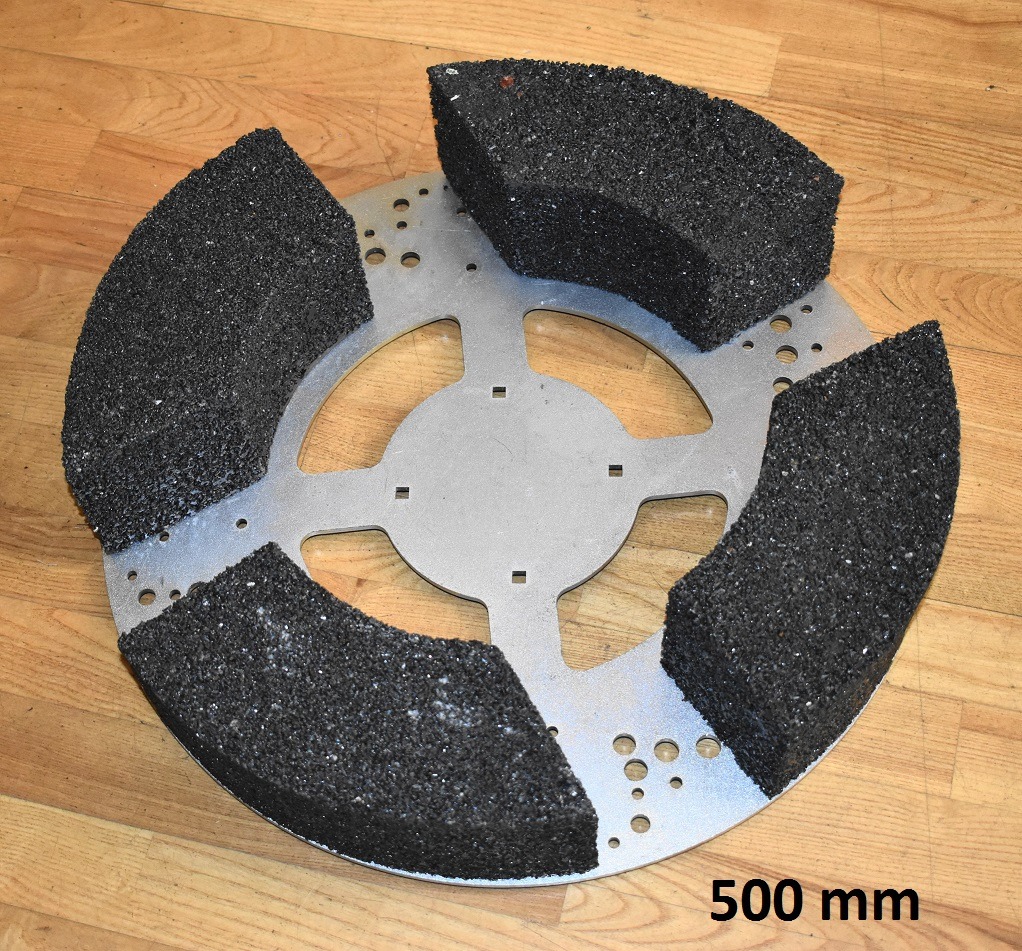Metalowa tarcza szlifierska o średnicy 500 mm z przymocowanymi segmentami ściernymi z czarnego, porowatego materiału, leżąca na drewnianej podłodze.