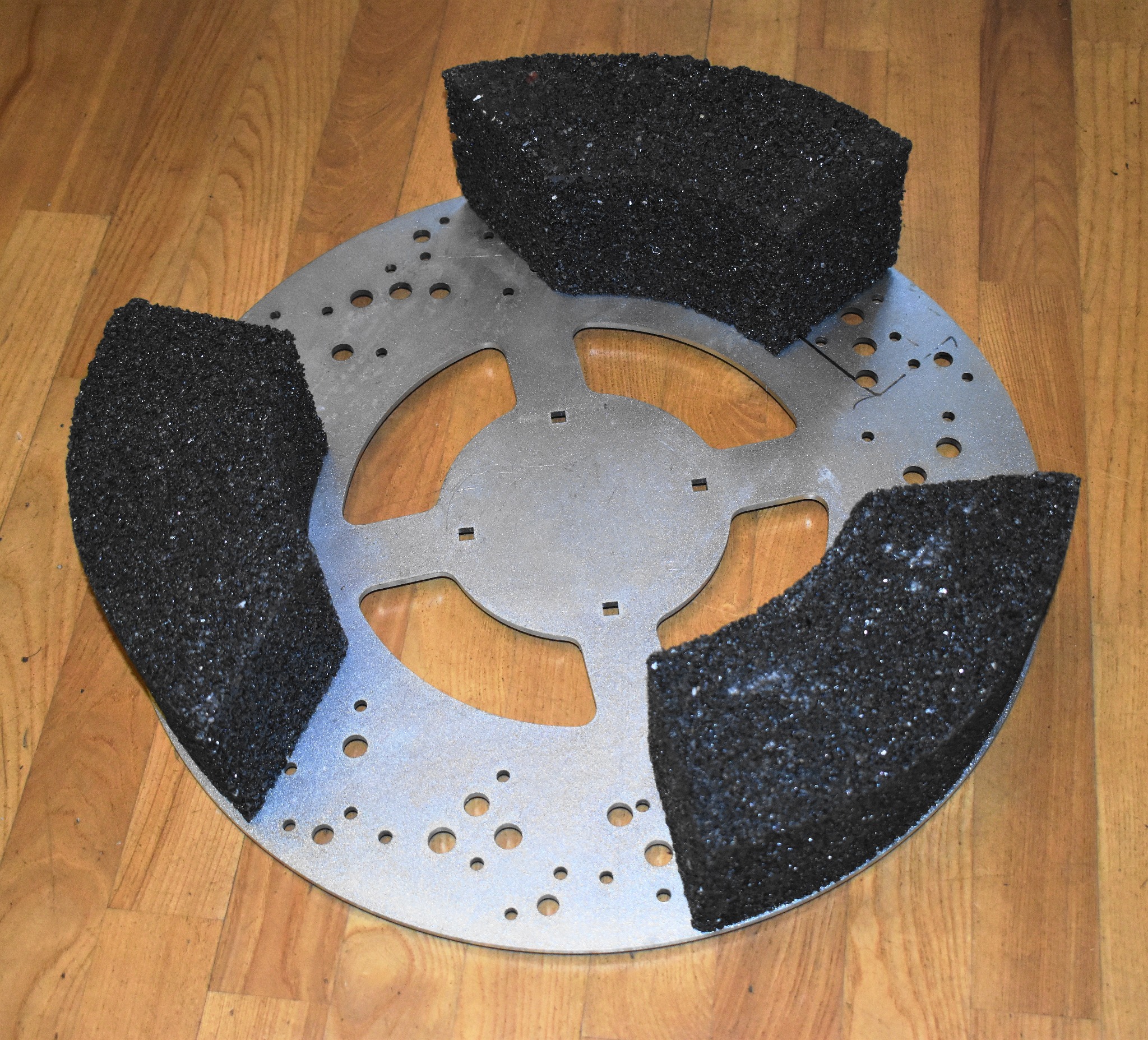 Metalowa tarcza z trzema czarnymi, porowatymi elementami przymocowanymi do jej krawędzi, leżąca na drewnianej podłodze.