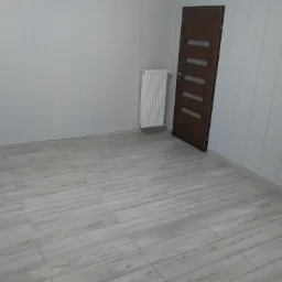 Puste pomieszczenie z panelami podłogowymi w odcieniach szarości, biały grzejnik żeberkowy i ciemne drzwi z przeszkleniami.