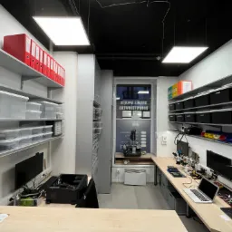 Wnętrze serwisu komputerowego z biurkami, komputerami, sprzętem diagnostycznym, półkami z pojemnikami i segregatorami z dokumentacją, widok z perspektywy osoby stojącej za biurkiem.