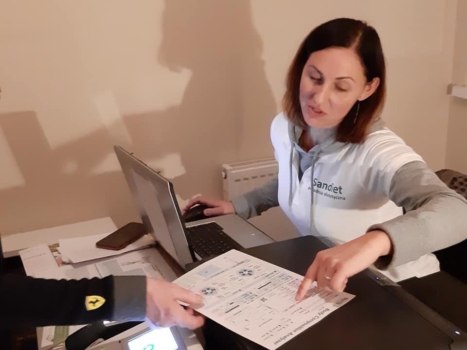 Kobieta w koszulce z logo 'Sandiet Akademia dietetyczna' analizuje wyniki analizy składu ciała na wydruku, wskazując palcem na konkretne dane; obok laptop i dokumenty.