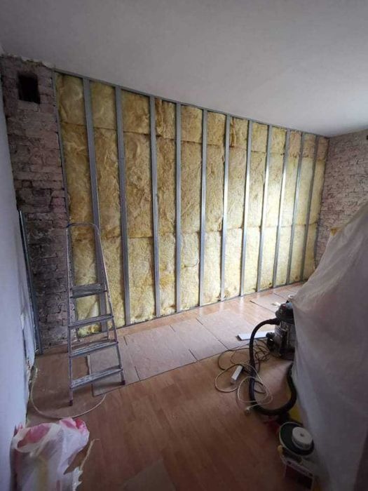 Ściana wewnętrzna w trakcie izolacji, widoczna konstrukcja metalowa z wypełnieniem z wełny mineralnej, surowy mur ceglany po lewej stronie, drabina i odkurzacz w pomieszczeniu.