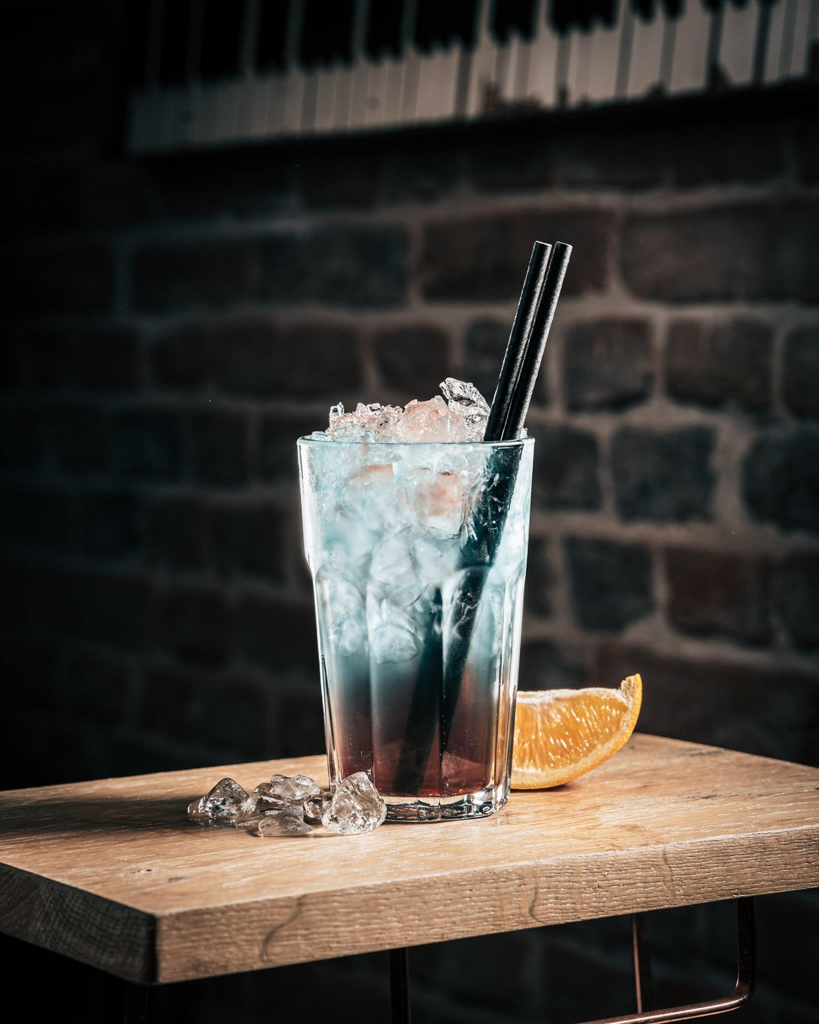 Warstwowy, błękitno-czerwony koktajl z lodem i dwoma czarnymi słomkami, ozdobiony plasterkiem pomarańczy, na drewnianym blacie z ceglanym tłem i klawiaturą w tle.