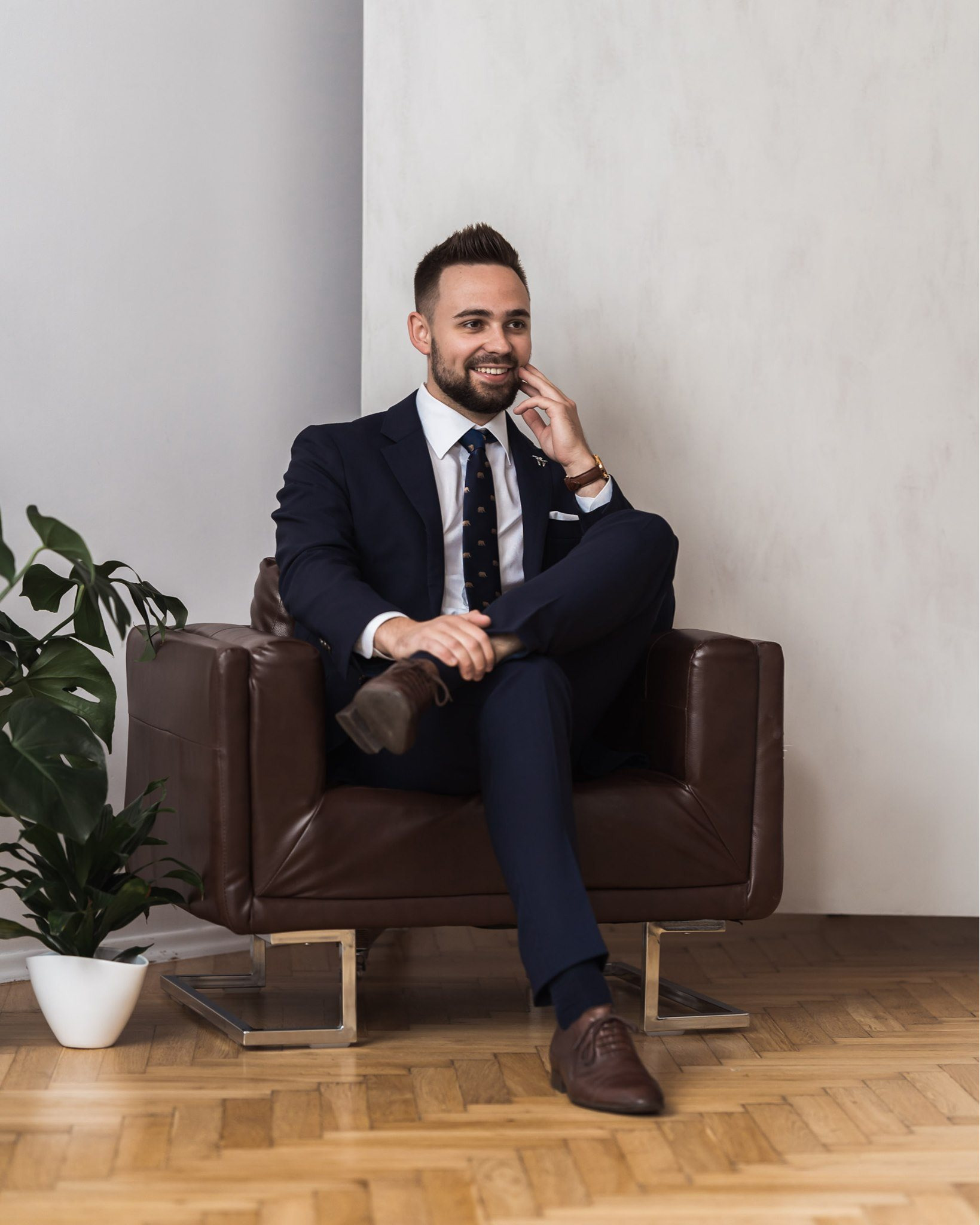 Mężczyzna w granatowym garniturze siedzi w skórzanym fotelu, z nogą na nodze, uśmiecha się, w tle biała ściana i roślina doniczkowa.