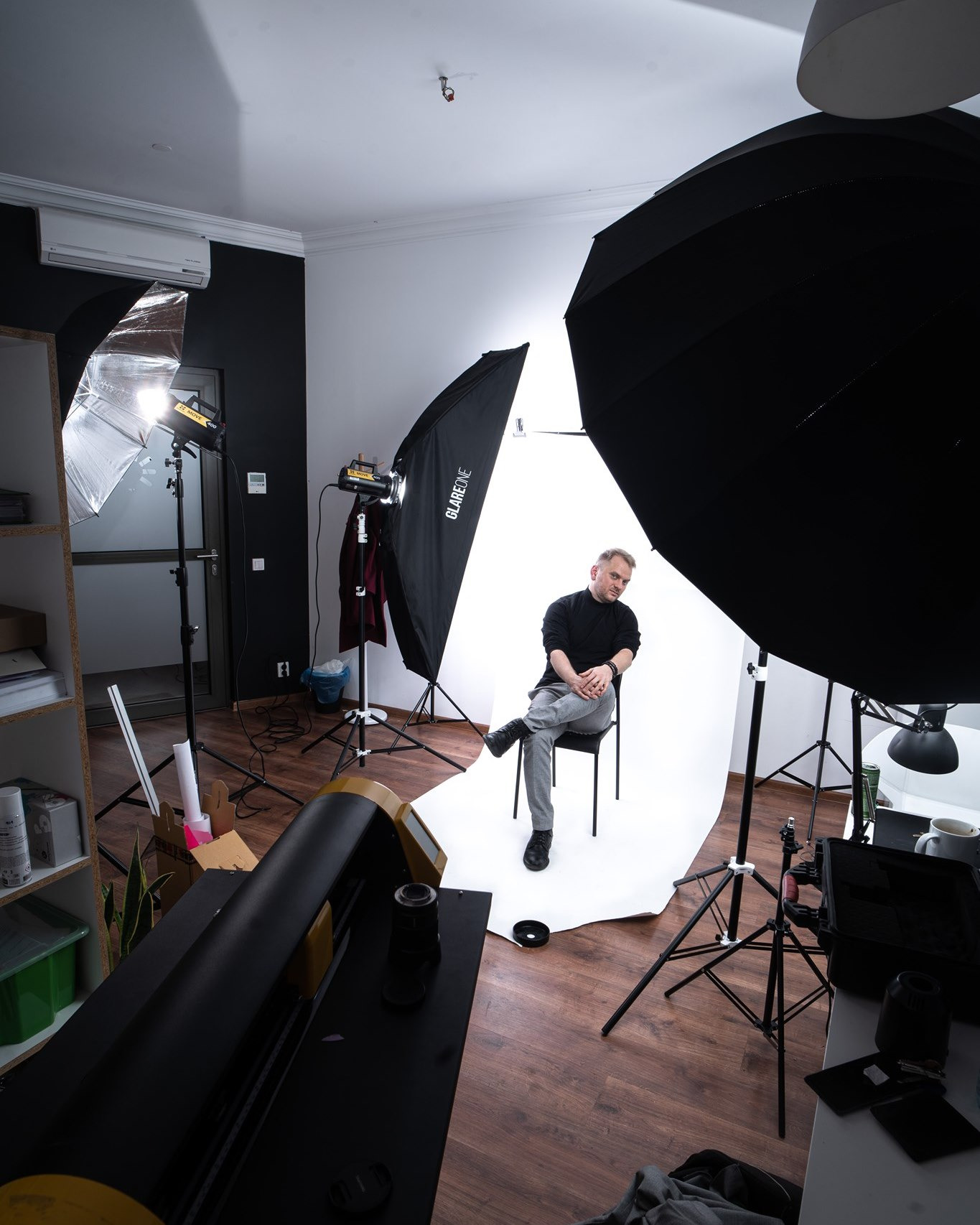 Mężczyzna pozuje na krześle w studio fotograficznym z oświetleniem i softboxami, widoczne wyposażenie studia i rekwizyty.