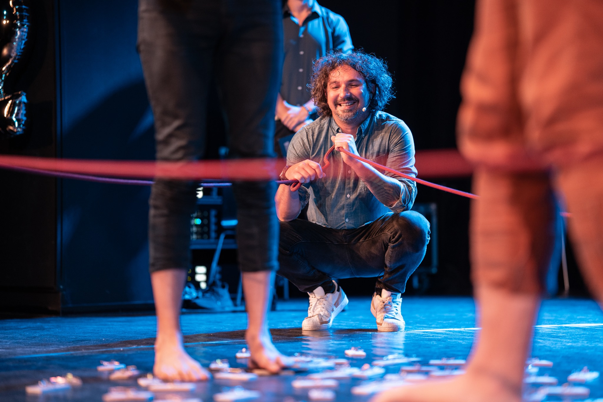 Uśmiechnięty mężczyzna w dżinsowej koszuli kuca na scenie, trzymając czerwoną linę, w tle inni ludzie i oświetlenie sceniczne, na podłodze leżą małe przedmioty.