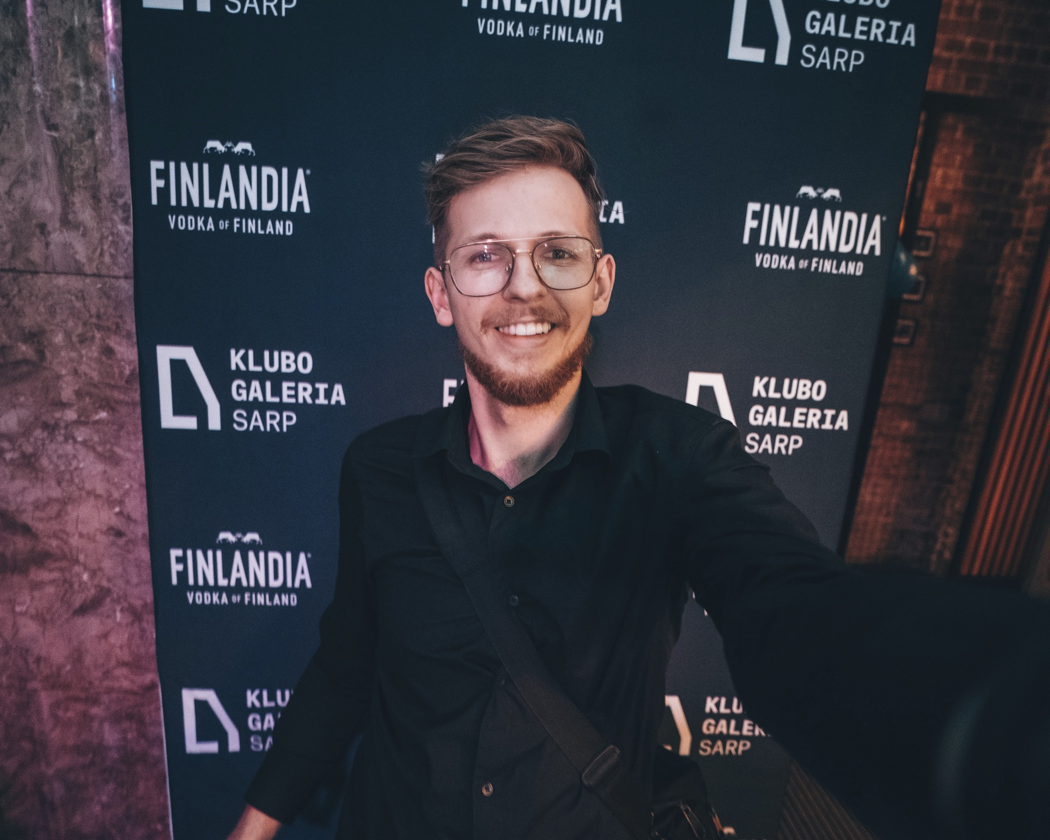 Uśmiechnięty mężczyzna w okularach, czarnej koszuli i torbie na ramieniu pozuje na tle ściany z logotypami Finlandia i Klubo Galeria Sarp.
