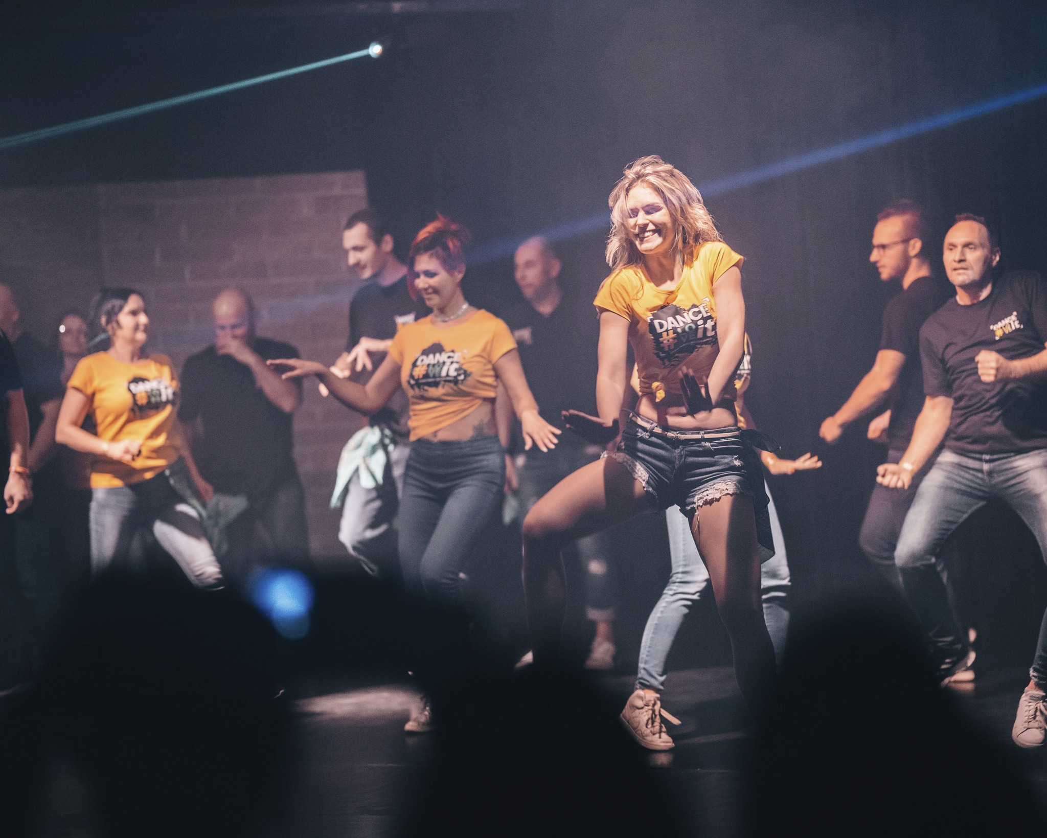 Grupa tancerzy w żółtych koszulkach z logo 'Dance #toit' na scenie podczas występu eventowego, oświetlona laserowym światłem.