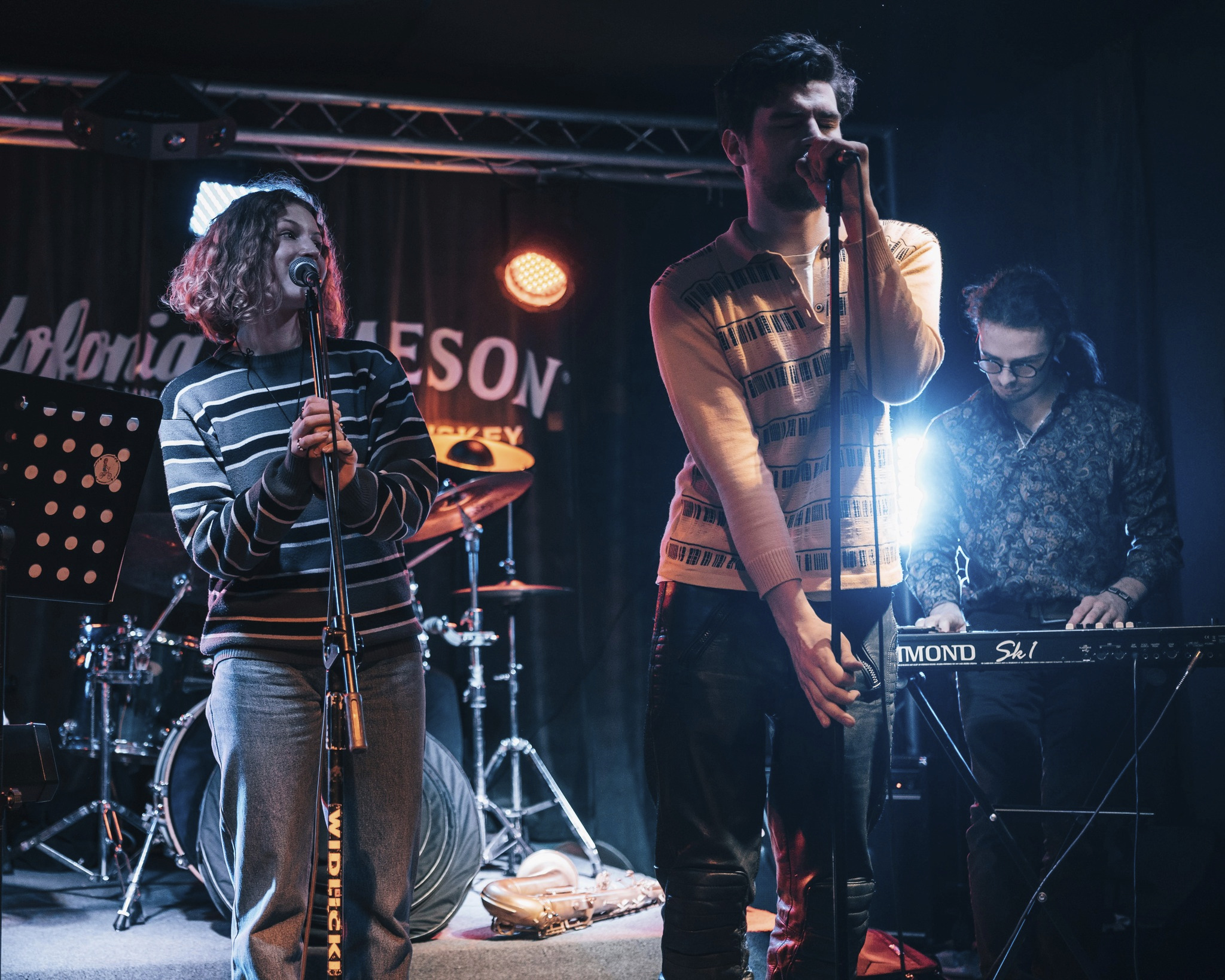 Zespół muzyczny w trakcie występu na scenie: wokalistka z mikrofonem, wokalista z mikrofonem i klawiszowiec grający na keyboardzie, w tle perkusja i saksofon.