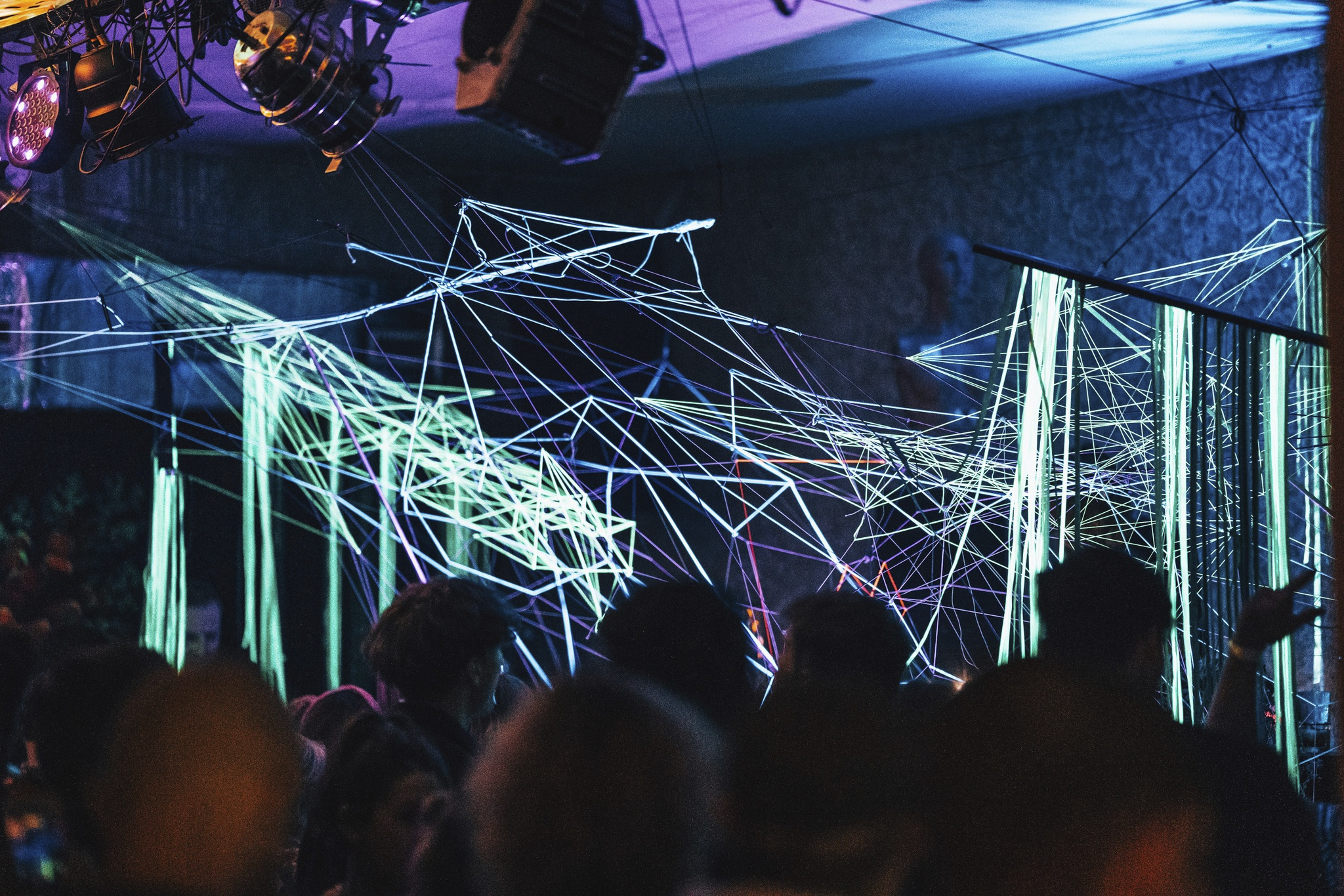 Widok z dołu na skomplikowaną instalację świetlną z geometrycznych linii w kolorach neonowej zieleni i fioletu, oświetloną reflektorami, z widocznym tłumem ludzi w tle.