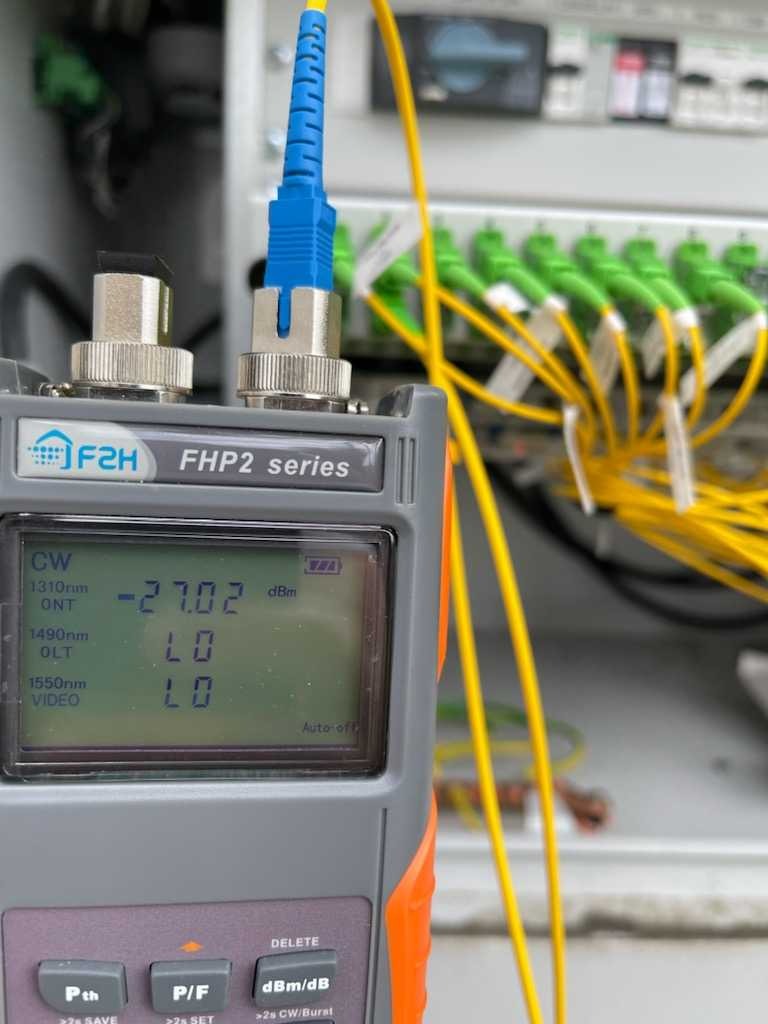 Miernik światłowodowy F2H FHP2 series podłączony do panelu światłowodowego z zielonymi złączkami, wskazujący wartość -27.02 dBm na wyświetlaczu.